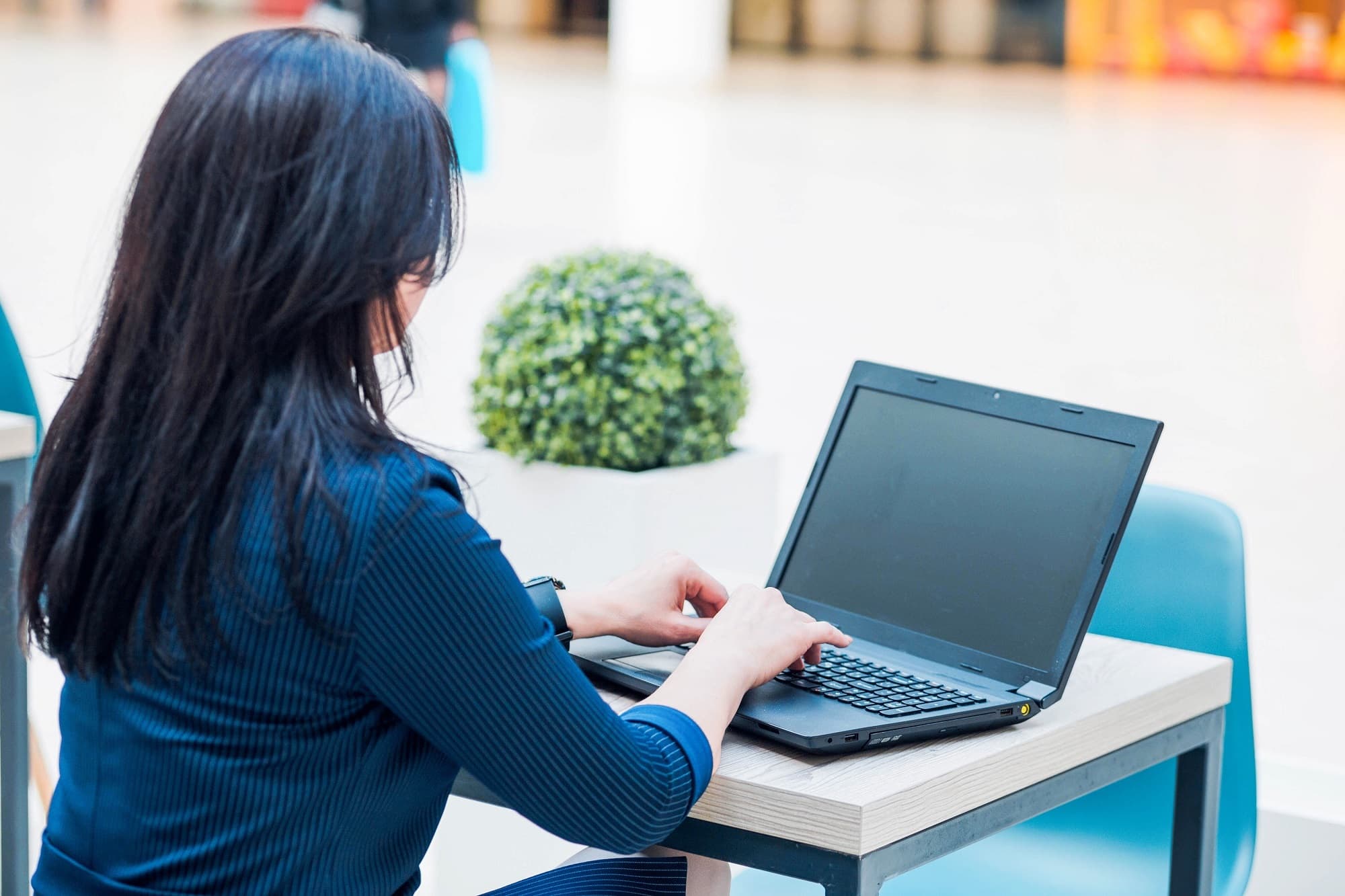 A business woman seated at a table in a public area using her laptop computer.