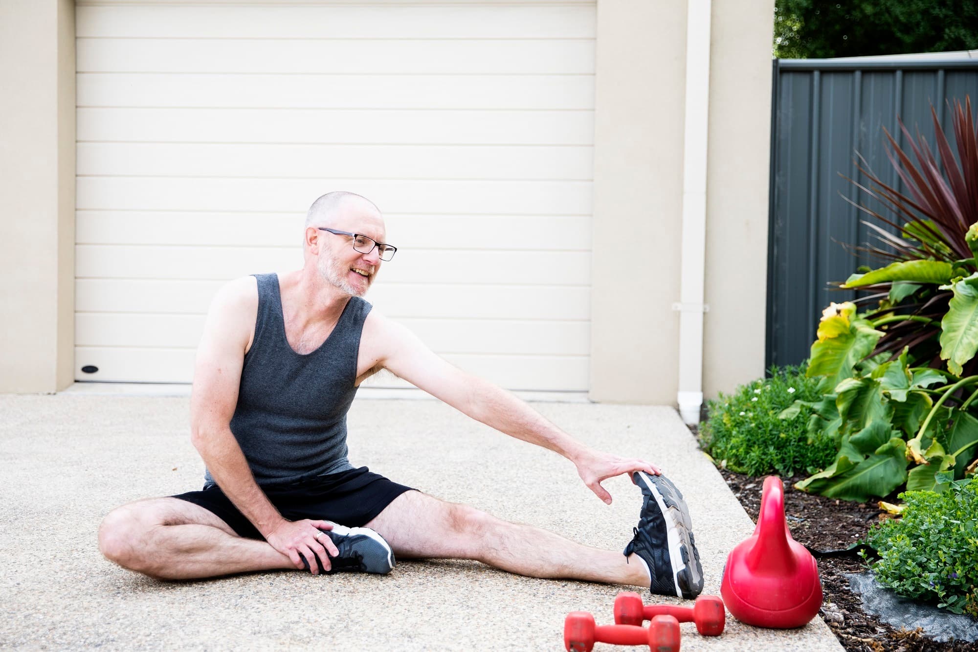 A senior man is seated in the driveway stretching out one leg.