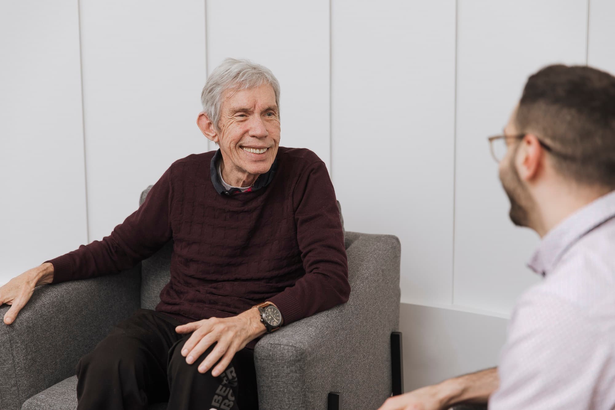 A senior man seated in a chair talking to another man.