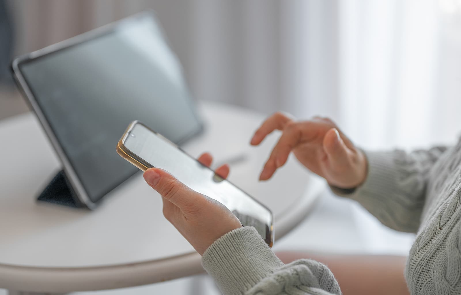a close up of hands holding and scrolling through a mobile with a tablet seated on a table next to them.