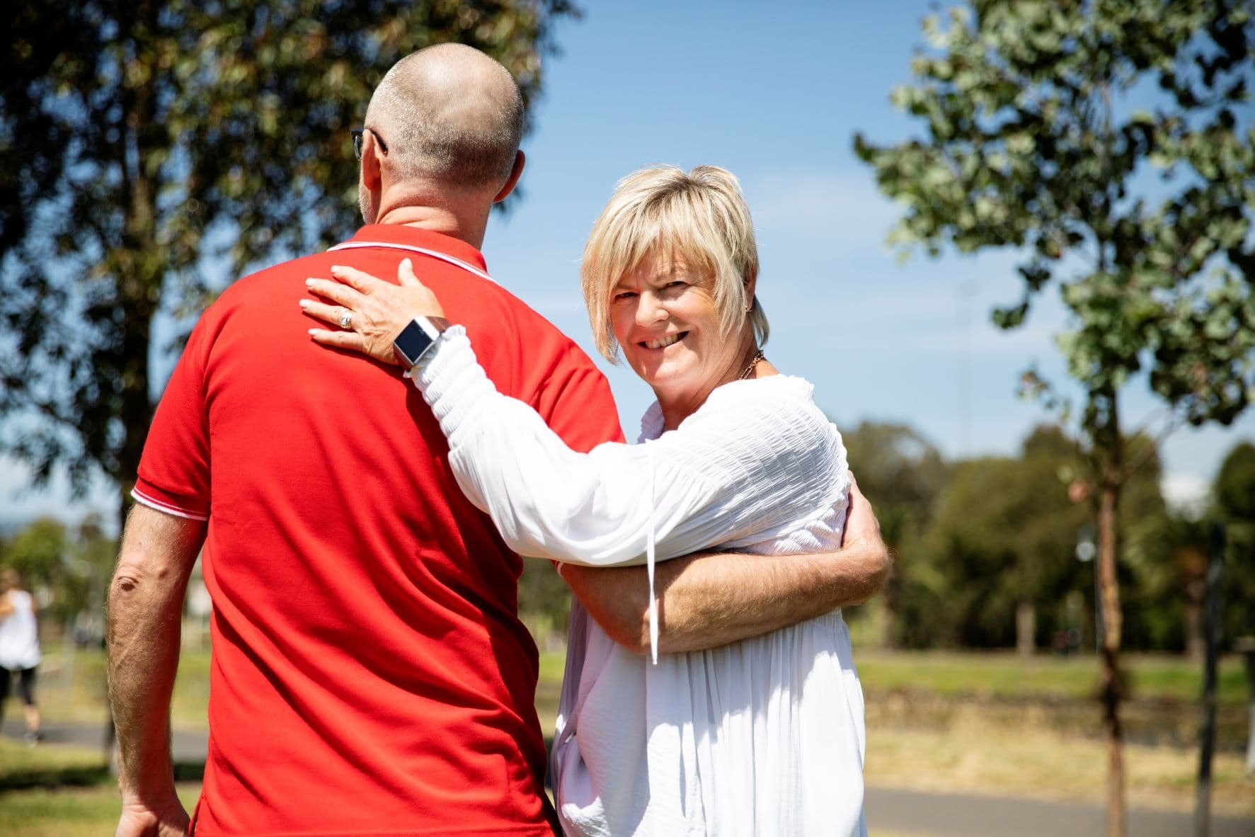 A man in red t-shirt and a woman in white shirt standing together with woman looking back, facing and smiling at the camera