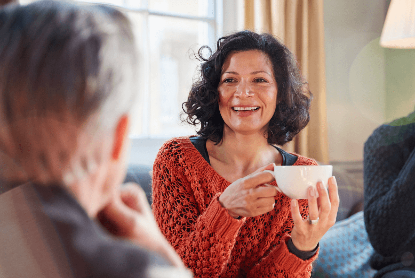 A woman holding a cup of tea seated on a couch talking to a man.