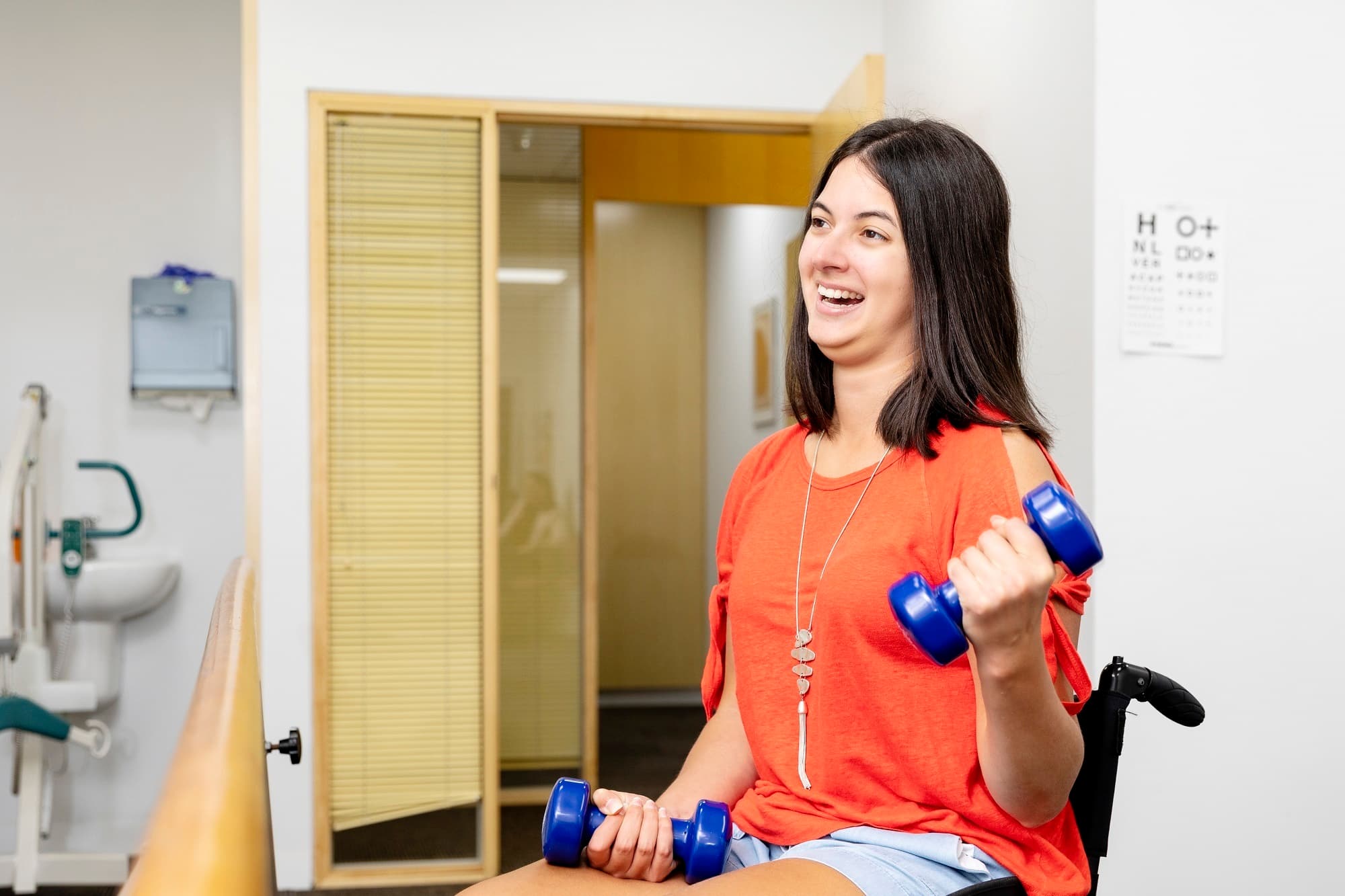 A woman with multiple sclerosis exercising with two small blue hand weights while seated in a wheelchair.
