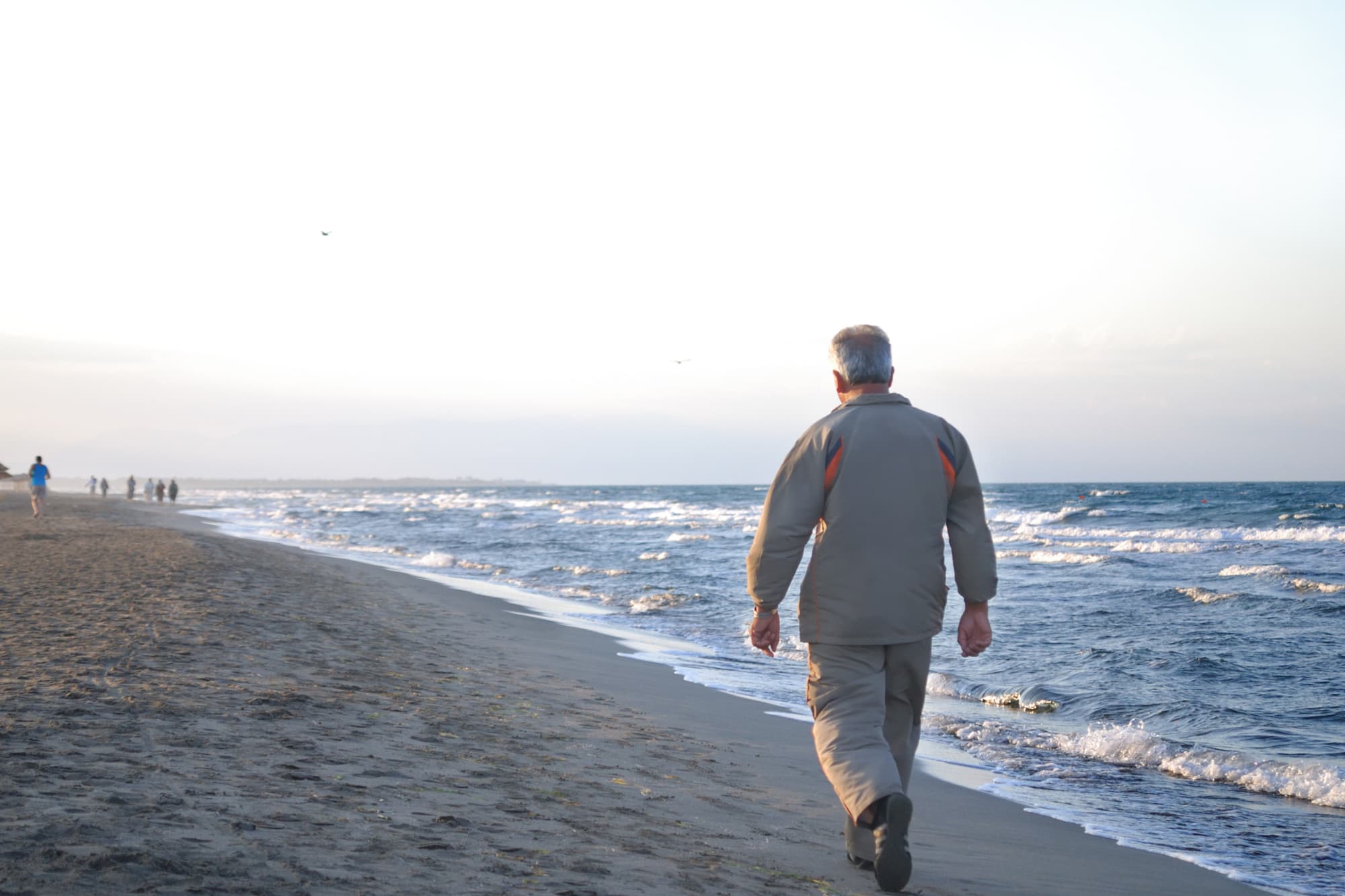 A rear view of a man wearing a beige tracksuit walking along the beach.